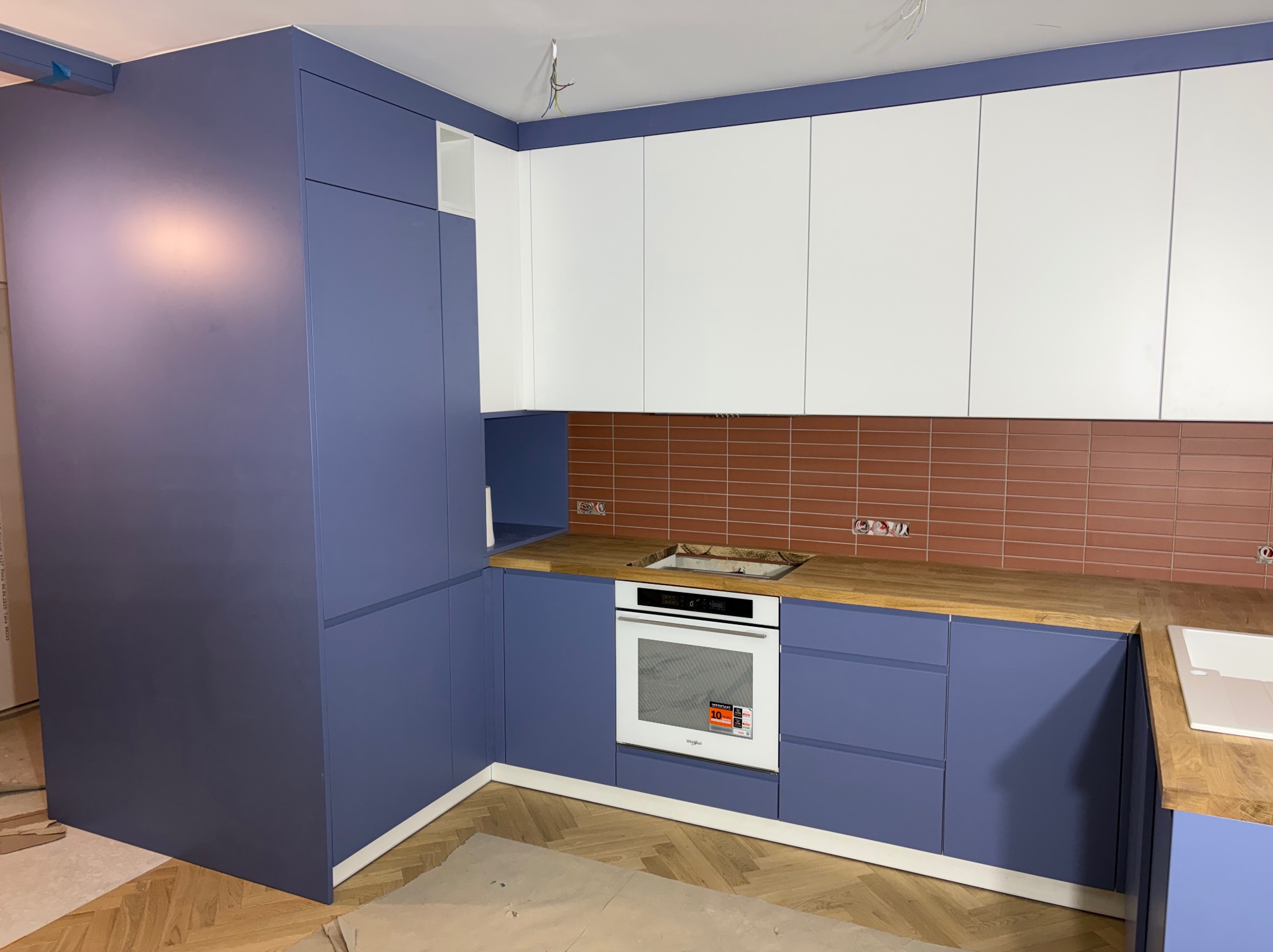 Nowoczesna kuchnia z zabudową w odcieniach bieli i fioletu, drewniany blat, brązowe płytki na ścianie. Widoczny piekarnik i fragment parkietu na podłodze.