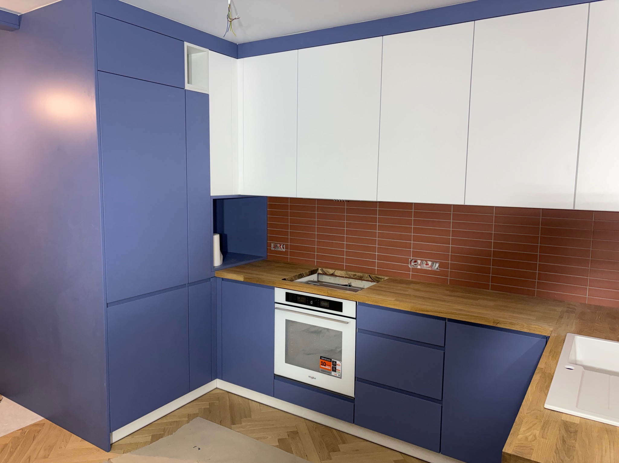 Nowoczesna kuchnia w odcieniach bieli i granatu z drewnianym blatem i ceglaną ścianą. Widoczny piekarnik i zlew. Podłoga wyłożona parkietem w jodełkę.