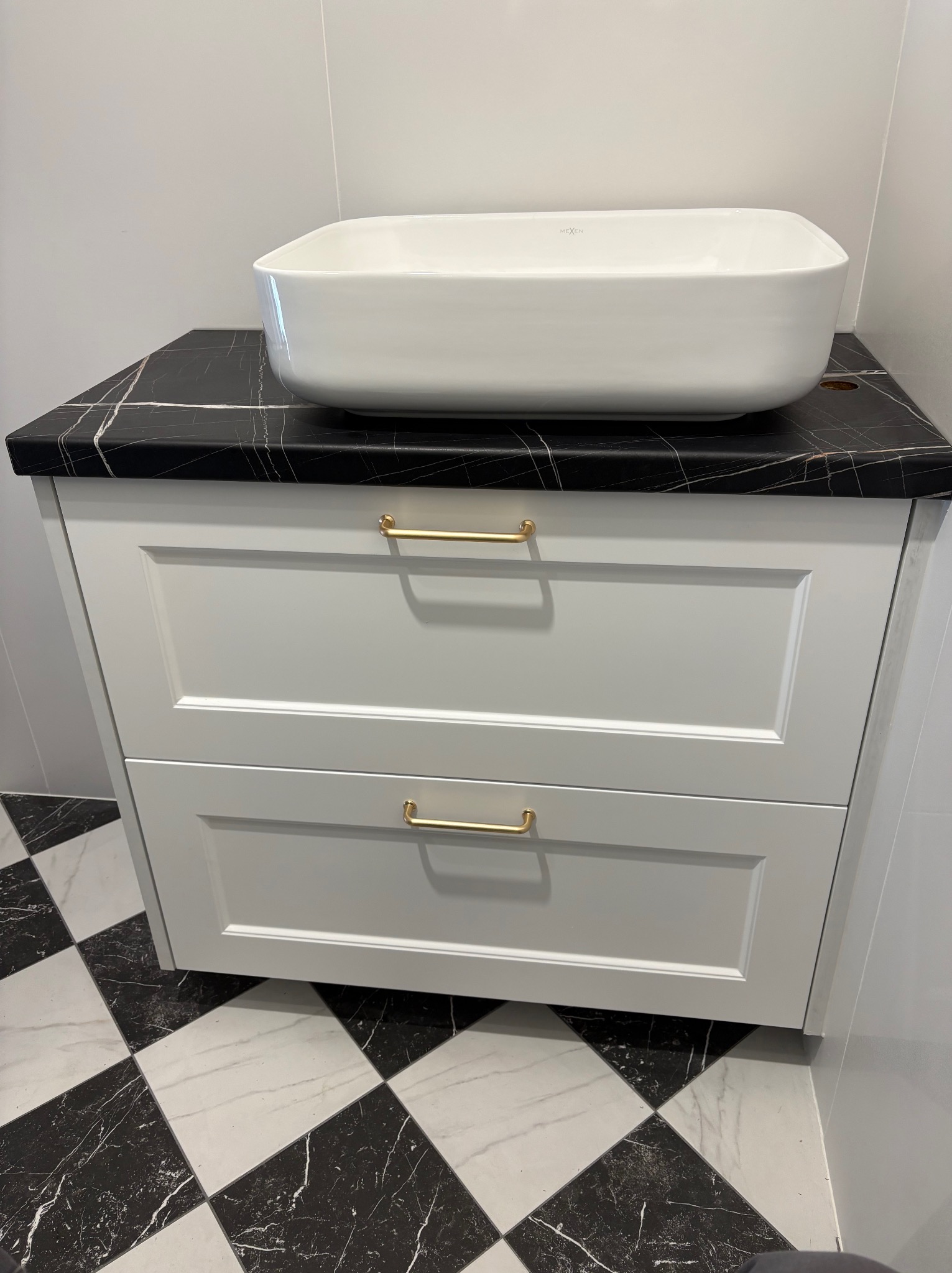 Biała szafka łazienkowa z dwoma szufladami i blatem imitującym czarny marmur, na której stoi prostokątna umywalka. Złote uchwyty. Podłoga w czarno-białą szachownicę.