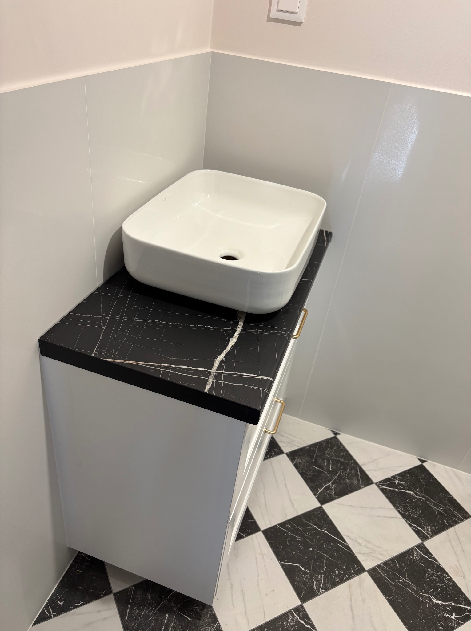 Biała, kwadratowa umywalka nablatowa na szafce z blatem imitującym czarny marmur. Złote uchwyty. W tle białe płytki i podłoga w czarno-białą szachownicę.