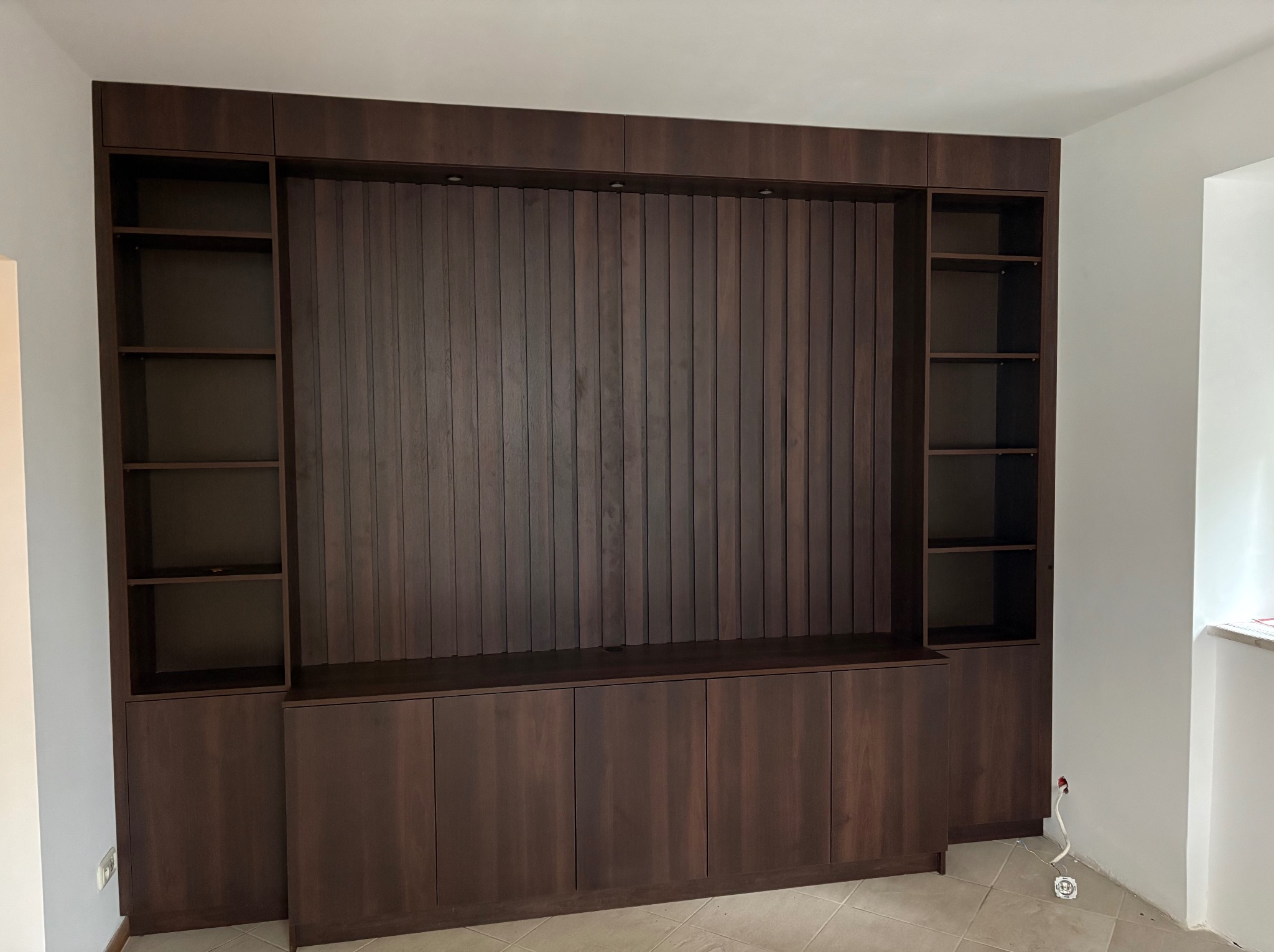 Ciemnobrązowa zabudowa meblowa z ryflowanym panelem ściennym i otwartymi półkami po bokach, oświetlona punktowo, na tle jasnych ścian.