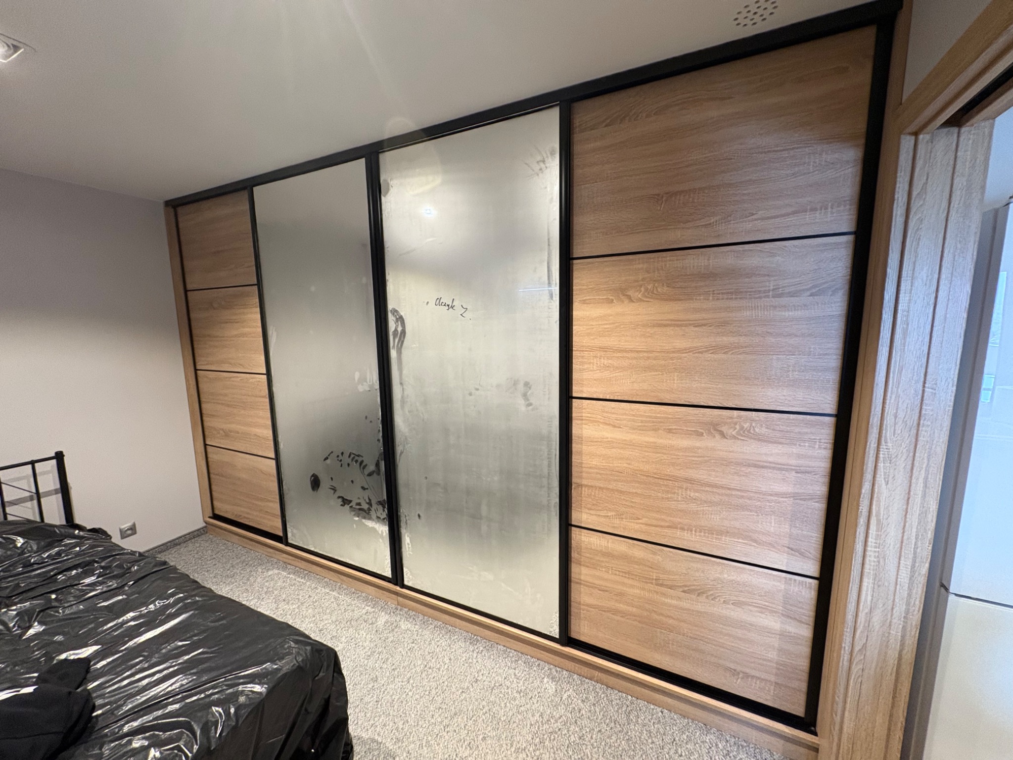 Szafa przesuwna z drewnianymi panelami i matowymi szybami, czarne obramowanie, widoczna w sypialni z łóżkiem przykrytym czarną folią. Nietypowe wykończenie szyb.