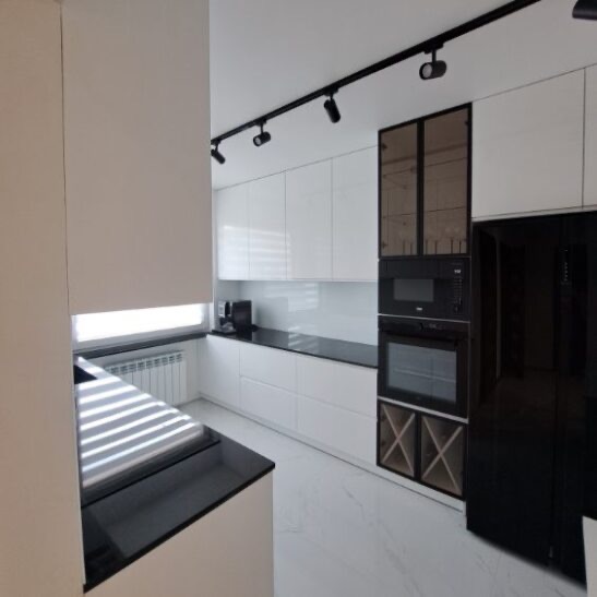 Biała kuchnia z czarnym blatem i sprzętem AGD, oświetlenie szynowe, zabudowa pod sufit z przeszklonymi szafkami. Nowoczesny design, minimalistyczny styl.
