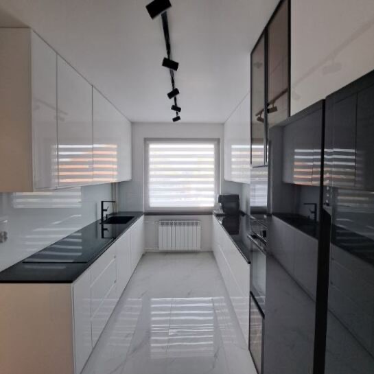 Nowoczesna, wąska kuchnia z białymi szafkami i czarnym blatem, oświetlona reflektorami. Okno z żaluzjami w tle. Błyszcząca podłoga odbija światło.