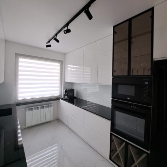 Biała, nowoczesna kuchnia z czarnym blatem i oświetleniem szynowym. Zabudowa z piekarnikiem i mikrofalówką, przeszklona szafka wisząca. Minimalistyczny design.