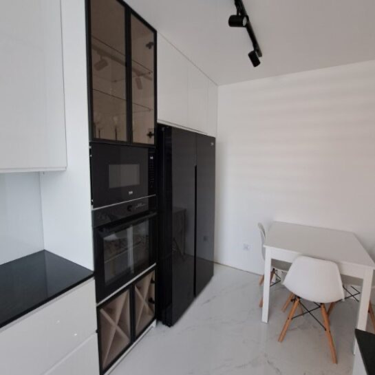 Biała kuchnia z czarnymi akcentami: zabudowa z piekarnikiem, mikrofalówką i szafką na wino. Obok czarna lodówka side-by-side i biały stół z krzesłami.