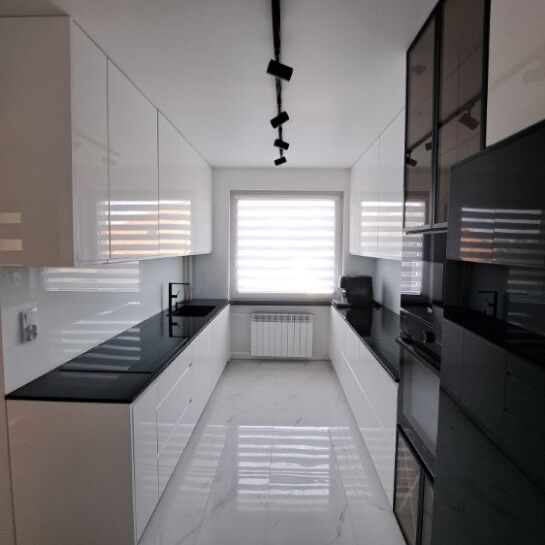 Nowoczesna, wąska kuchnia z białymi, błyszczącymi szafkami i czarnym blatem. Widoczne oświetlenie punktowe i marmurowa podłoga. Minimalistyczny design.
