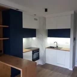 Nowoczesna kuchnia z granatowymi i białymi szafkami, blatem z jasnego drewna i marmurową ścianą, oświetlona punktowo, z drewnianą wyspą kuchenną.