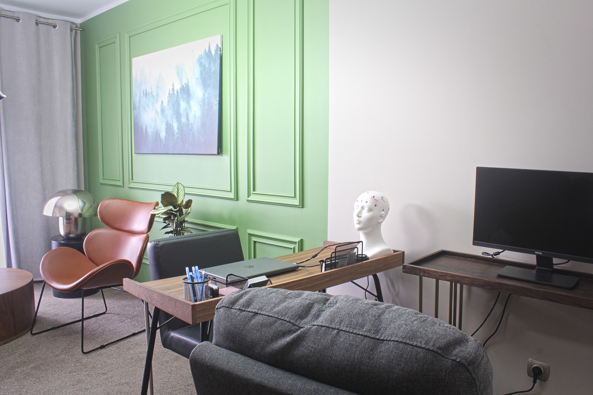 Gabinet z biurkiem, laptopem, manekinem głowy z elektrodami EEG, krzesłem i obrazem górskiego lasu we mgle na ścianie.