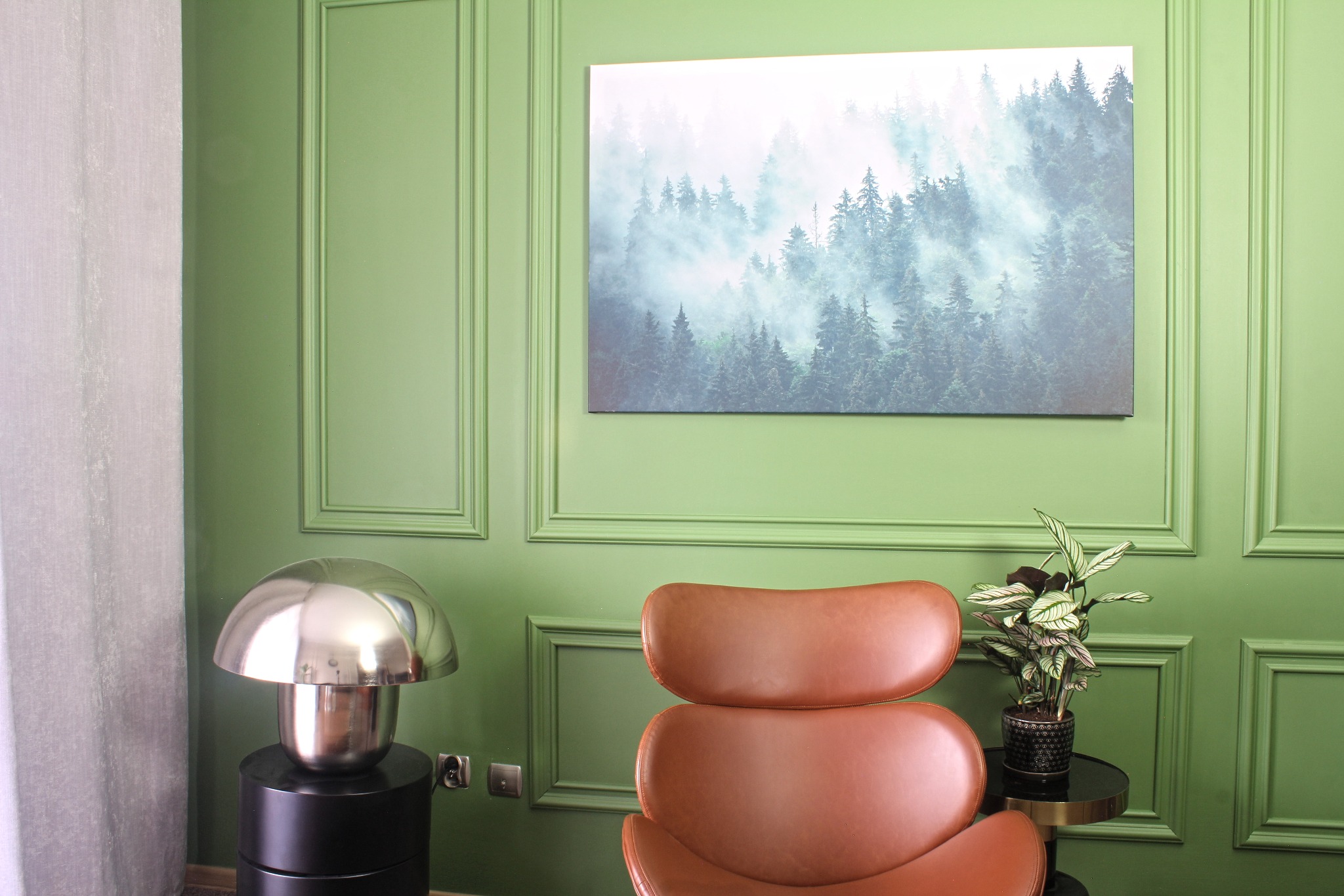 Pokój z pistacjowymi ścianami w klasycznej sztukaterii, brązowy skórzany fotel, metalowa lampa w stylu retro, stolik z rośliną doniczkową i obraz przedstawiający zamglony las.