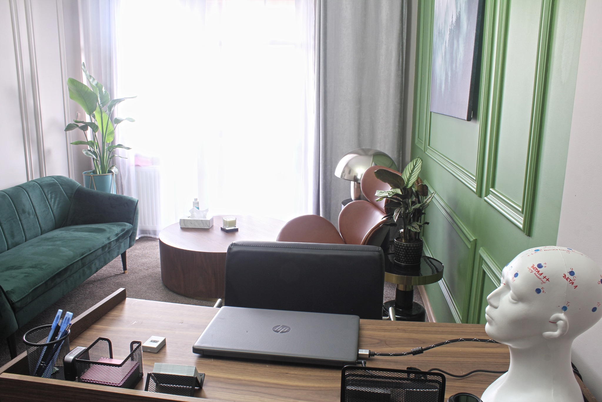 Gabinet z zieloną sofą, okrągłym stolikiem, laptopem na biurku i modelem głowy z zaznaczonymi punktami pomiarowymi, sugerujący przestrzeń do konsultacji.