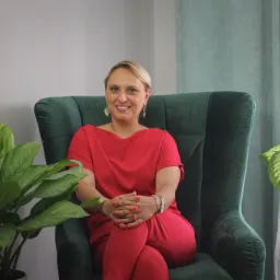 Kobieta w czerwonym komplecie siedzi w zielonym fotelu, uśmiechając się; obok rośliny doniczkowe, w tle zasłony.