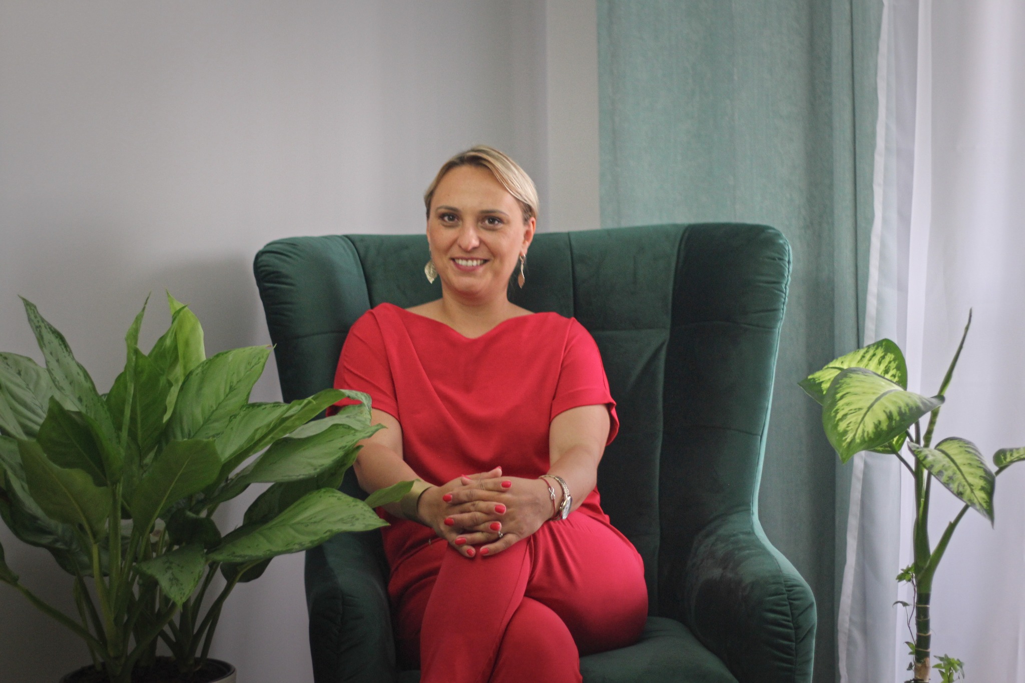 Kobieta w czerwonym komplecie siedzi w zielonym fotelu, uśmiechając się; obok rośliny doniczkowe, w tle zasłony.