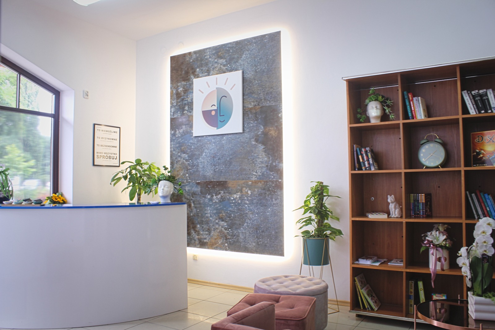 Jasne wnętrze gabinetu z recepcją, dekoracyjnym panelem ściennym z podświetleniem LED, regałem z książkami i roślinami doniczkowymi.