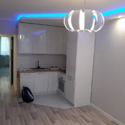 Nowoczesna, biała kuchnia z niebieskim oświetleniem LED w Toruniu, z wiszącą lampą o okrągłym kształcie i widocznym fragmentem podłogi oraz torbą sportową leżącą na podłodze.