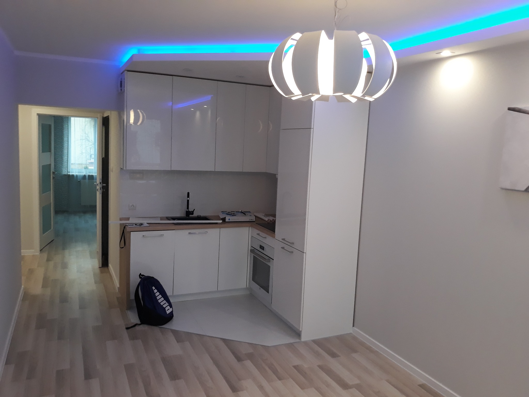 Nowoczesna, biała kuchnia z niebieskim oświetleniem LED w Toruniu, z wiszącą lampą o okrągłym kształcie i widocznym fragmentem podłogi oraz torbą sportową leżącą na podłodze.