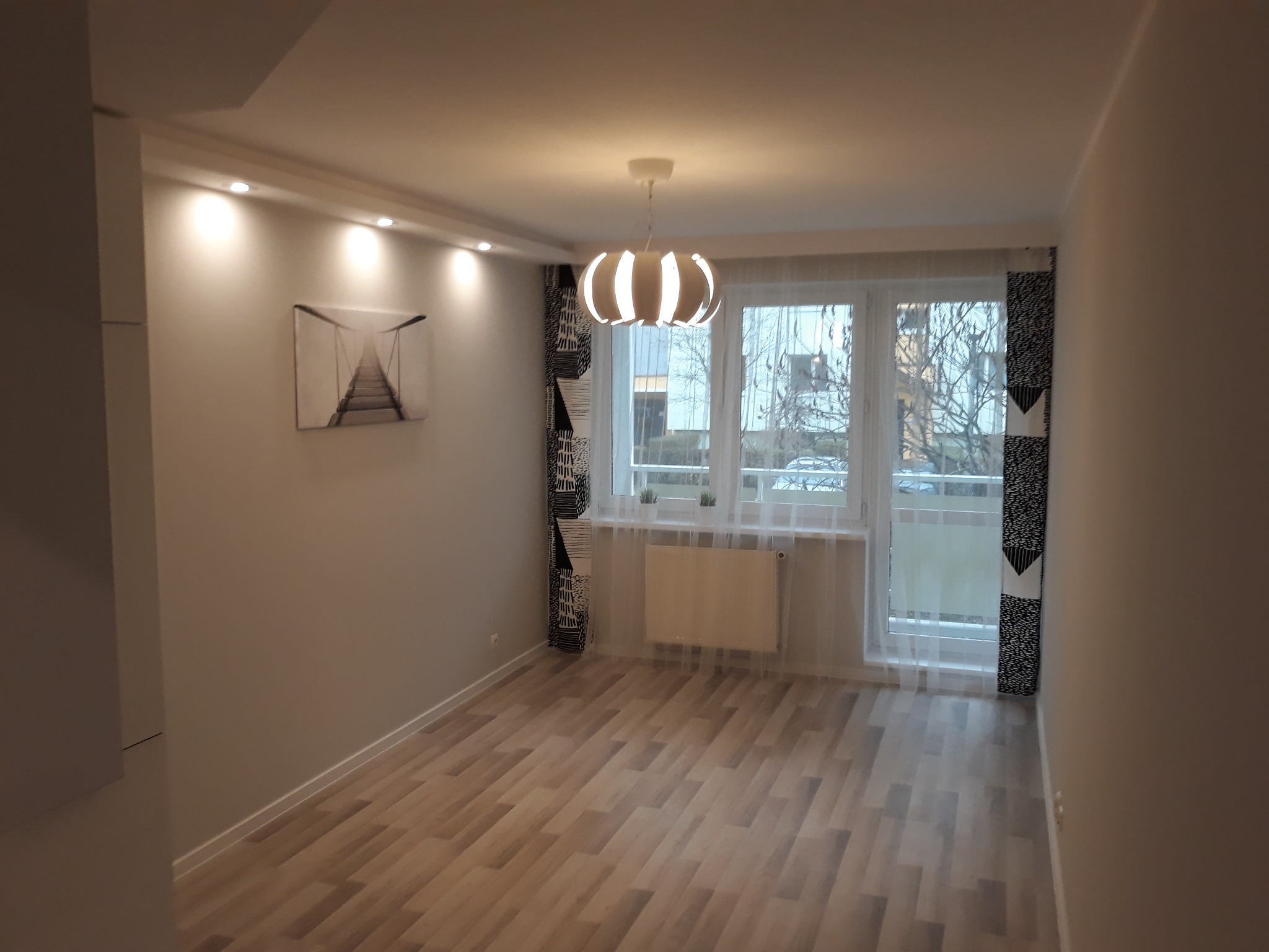 Pusty pokój po remoncie z panelami podłogowymi, białymi ścianami, grzejnikiem pod oknem i nowoczesną lampą wiszącą.