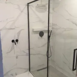 Nowoczesna kabina prysznicowa z czarną ramą i deszczownicą, białe płytki imitujące marmur na ścianach, czarna armatura, odpływ liniowy w podłodze wyłożonej szarą płytką.