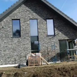 Dom jednorodzinny z elewacją z szarej cegły klinkierowej, widoczne rusztowania i przygotowany teren wokół budynku.