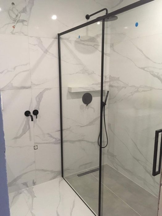 Nowoczesna kabina prysznicowa z czarną ramą i deszczownicą, białe płytki imitujące marmur na ścianach, czarna armatura, odpływ liniowy w podłodze wyłożonej szarą płytką.