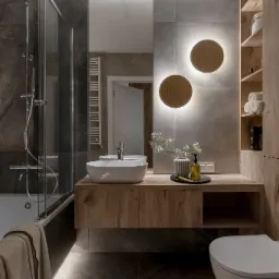 Nowoczesna łazienka z marmurowymi płytkami, drewnianą szafką pod umywalką, okrągłymi lampami ściennymi i minimalistycznym wyposażeniem.