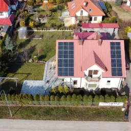 Widok z góry na dom jednorodzinny z czerwoną dachówką i panelami fotowoltaicznymi na dachu, otoczony zadbanym ogrodem i innymi domami.