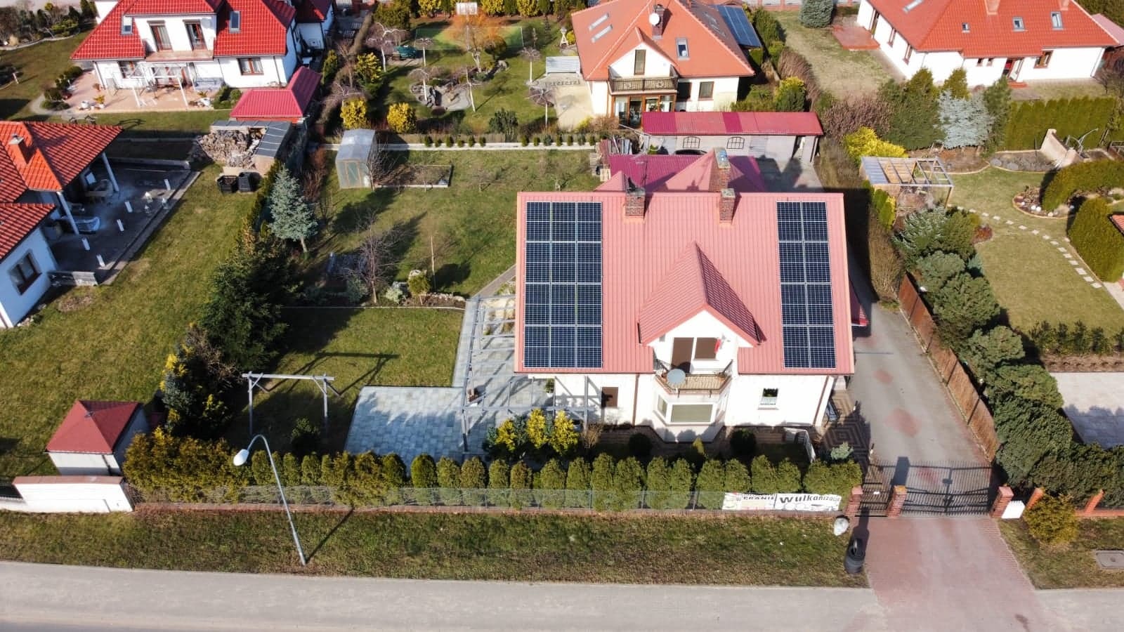 Widok z góry na dom jednorodzinny z czerwoną dachówką i panelami fotowoltaicznymi na dachu, otoczony zadbanym ogrodem i innymi domami.