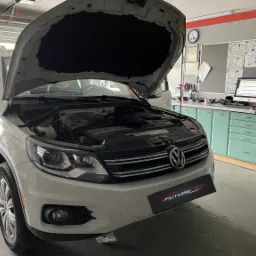 Biały Volkswagen Tiguan z otwartą maską w warsztacie, widoczne schematy na tablicy i komputer diagnostyczny.