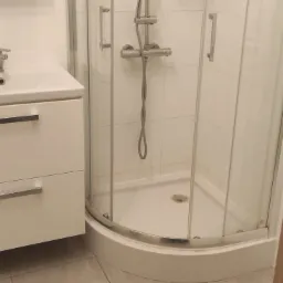 Jasna łazienka z kabiną prysznicową z zaokrąglonymi szybami i białą szafką pod umywalką z dwoma szufladami oraz chromowanymi uchwytami.