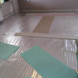 Przygotowanie podłoża pod panele podłogowe: folia izolacyjna i zielone płyty izolacyjne rozłożone na nierównej podłodze w trakcie remontu.