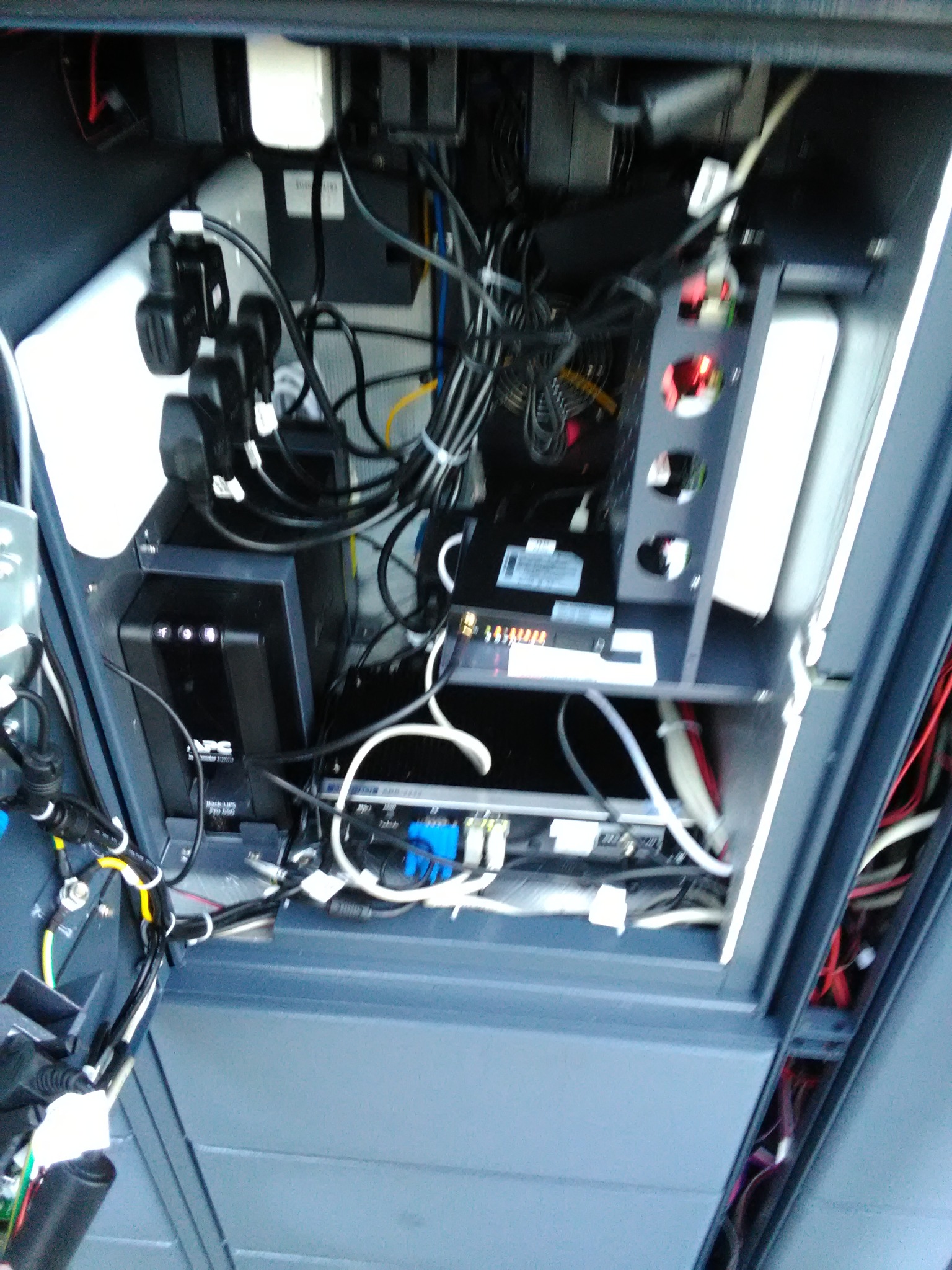 Wnętrze szafy telekomunikacyjnej z zainstalowanym zasilaczem awaryjnym APC, routerem i licznymi kablami, widok z dołu.