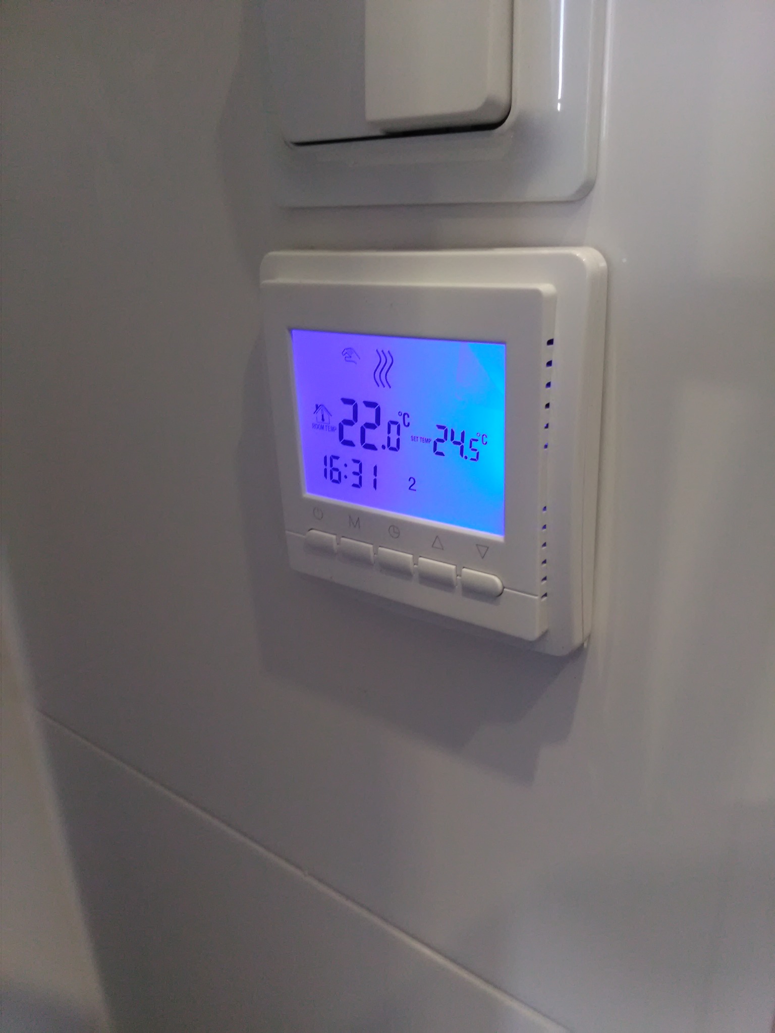 Biały, cyfrowy termostat pokojowy z podświetlanym na niebiesko wyświetlaczem, zamontowany na białej ścianie, obok włącznika światła.