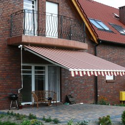 Przedsiębiorstwo Usługowo Handlowo Produkcyjne EUROL Małgorzata Czarnotta - Czerwono-biała markiza w paski rozłożona nad tarasem z kostki brukowej przy ceglanym domu z balkonem z kutą balustradą.