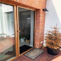 Przedsiębiorstwo Usługowo Handlowo Produkcyjne EUROL Małgorzata Czarnotta - Drewniana rama drzwi wejściowych z siatką ochronną, widoczna z zewnątrz na tle ceglanej ściany i białej elewacji, obok donica z uschniętym drzewkiem.