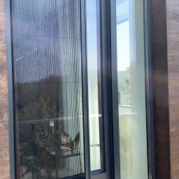 Przedsiębiorstwo Usługowo Handlowo Produkcyjne EUROL Małgorzata Czarnotta - Zasłonięte okno z moskitierą plisowaną w kolorze grafitowym, zamontowaną w nowoczesnym domu z elewacją imitującą drewno.