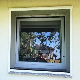 Przedsiębiorstwo Usługowo Handlowo Produkcyjne EUROL Małgorzata Czarnotta - Białe okno z zamontowaną moskitierą rolowaną, widoczne odbicie drzewa i osoby robiącej zdjęcie.