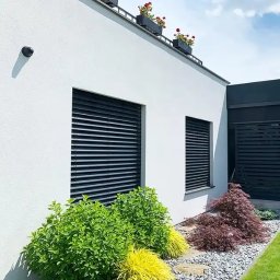 Przedsiębiorstwo Usługowo Handlowo Produkcyjne EUROL Małgorzata Czarnotta - Nowoczesny dom z białymi ścianami, czarnymi żaluzjami fasadowymi, roślinami w donicach na dachu i ozdobnym ogrodem z kamykami.
