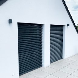 Przedsiębiorstwo Usługowo Handlowo Produkcyjne EUROL Małgorzata Czarnotta - Dwa okna zabezpieczone zewnętrznymi żaluzjami fasadowymi w kolorze grafitowym, zamontowane w białej ścianie budynku z ciemnym dachem, widoczne z poziomu tarasu.