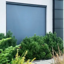 Zewnętrzna roleta w kolorze grafitowym na tle białej elewacji, zasłonięta, z widocznymi krzewami iglastymi w ogrodzie.