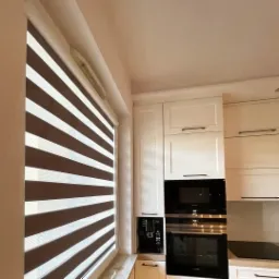 Zebra żaluzja w kolorze brązowo-białym zamontowana w oknie w kuchni w Opolu, z widocznymi białymi szafkami kuchennymi i sprzętem AGD w tle.