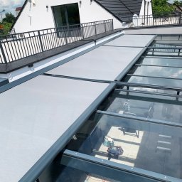 Przedsiębiorstwo Usługowo Handlowo Produkcyjne EUROL Małgorzata Czarnotta - Szare, rozsuwane osłony przeciwsłoneczne na szklanym dachu oranżerii, widok z zewnątrz na biały budynek z czarnym balkonem.