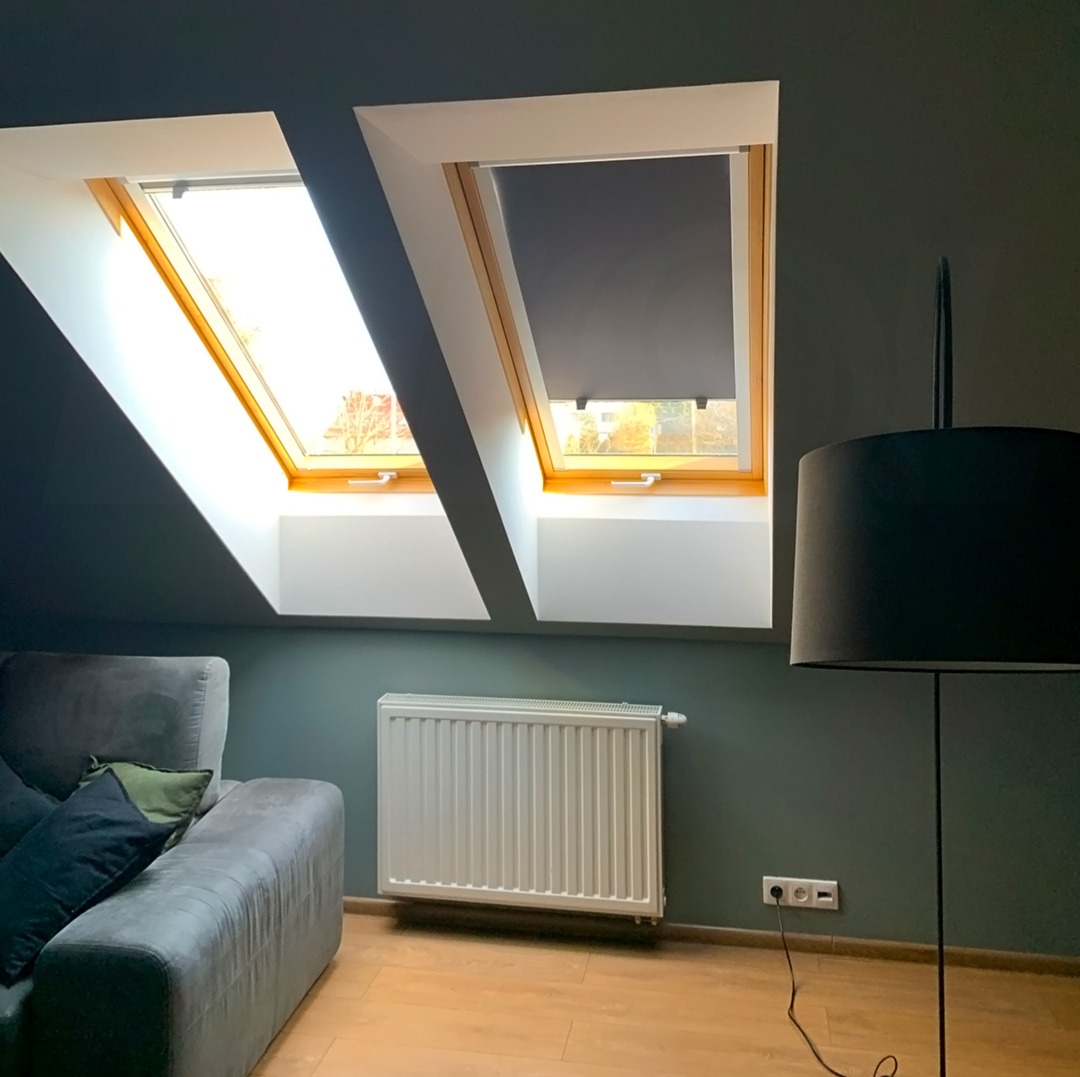 Dwa okna dachowe z drewnianą ramą, jedno z opuszczoną szarą roletą, w pokoju z szarą sofą i lampą.