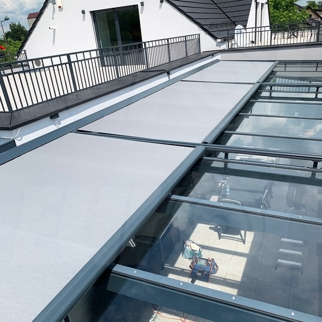 Szare, rozsuwane osłony przeciwsłoneczne na szklanym dachu oranżerii, widok z zewnątrz na biały budynek z czarnym balkonem.