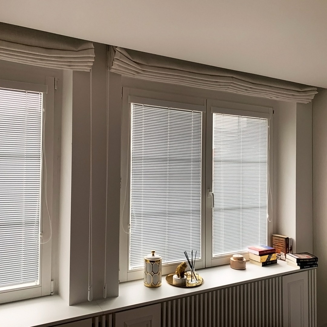 Trzy okna z białymi, poziomymi żaluzjami i szarymi roletami rzymskimi u góry, dekoracje na parapecie.