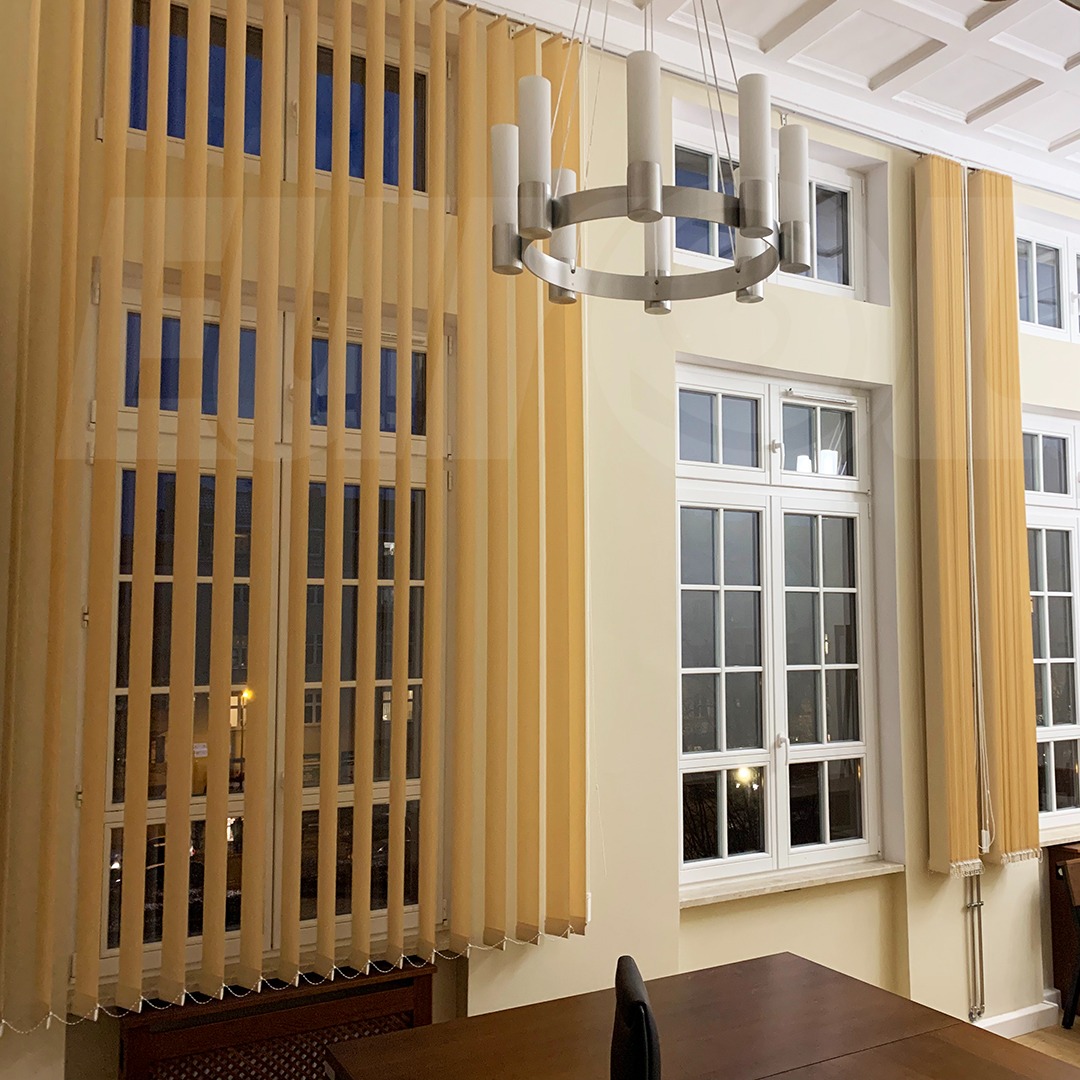 Beżowe żaluzje pionowe na oknach w jasnym pomieszczeniu z nowoczesnym żyrandolem.