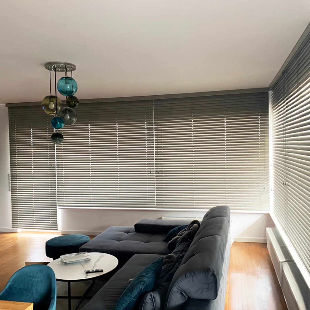 Szare poziome żaluzje w nowoczesnym salonie z szarą sofą i drewnianą podłogą, oświetlone lampą z kolorowymi kulami.