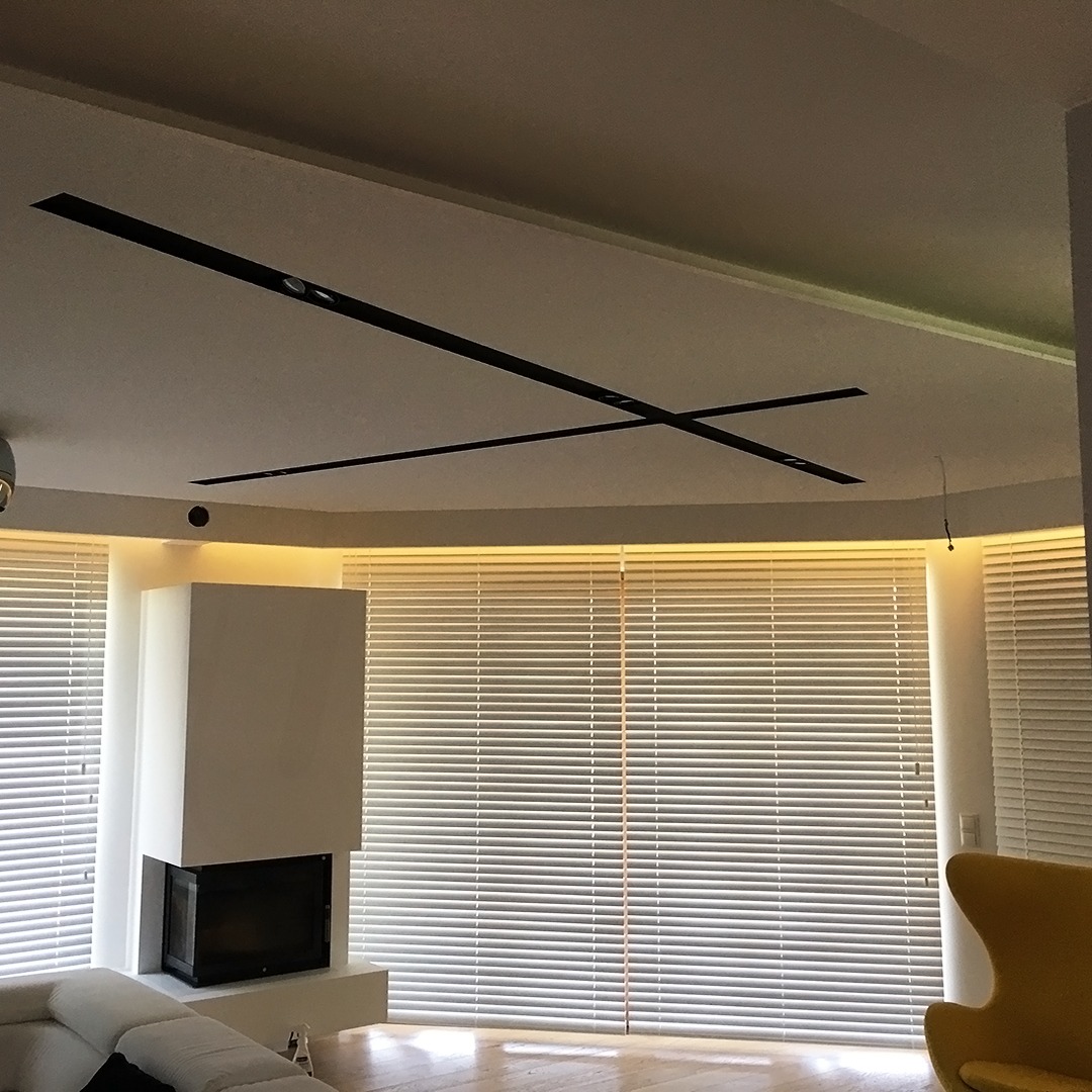 Jasne wnętrze z dużymi oknami osłoniętymi poziomymi żaluzjami w kolorze białym, widoczny kominek i fragment sofy.