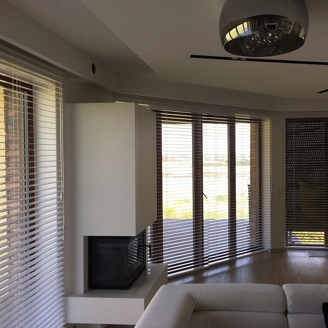 Jasne wnętrze z kominkiem i oknami wyposażonymi w poziome żaluzje drewniane w odcieniach brązu i bieli, zapewniające prywatność i kontrolę światła.