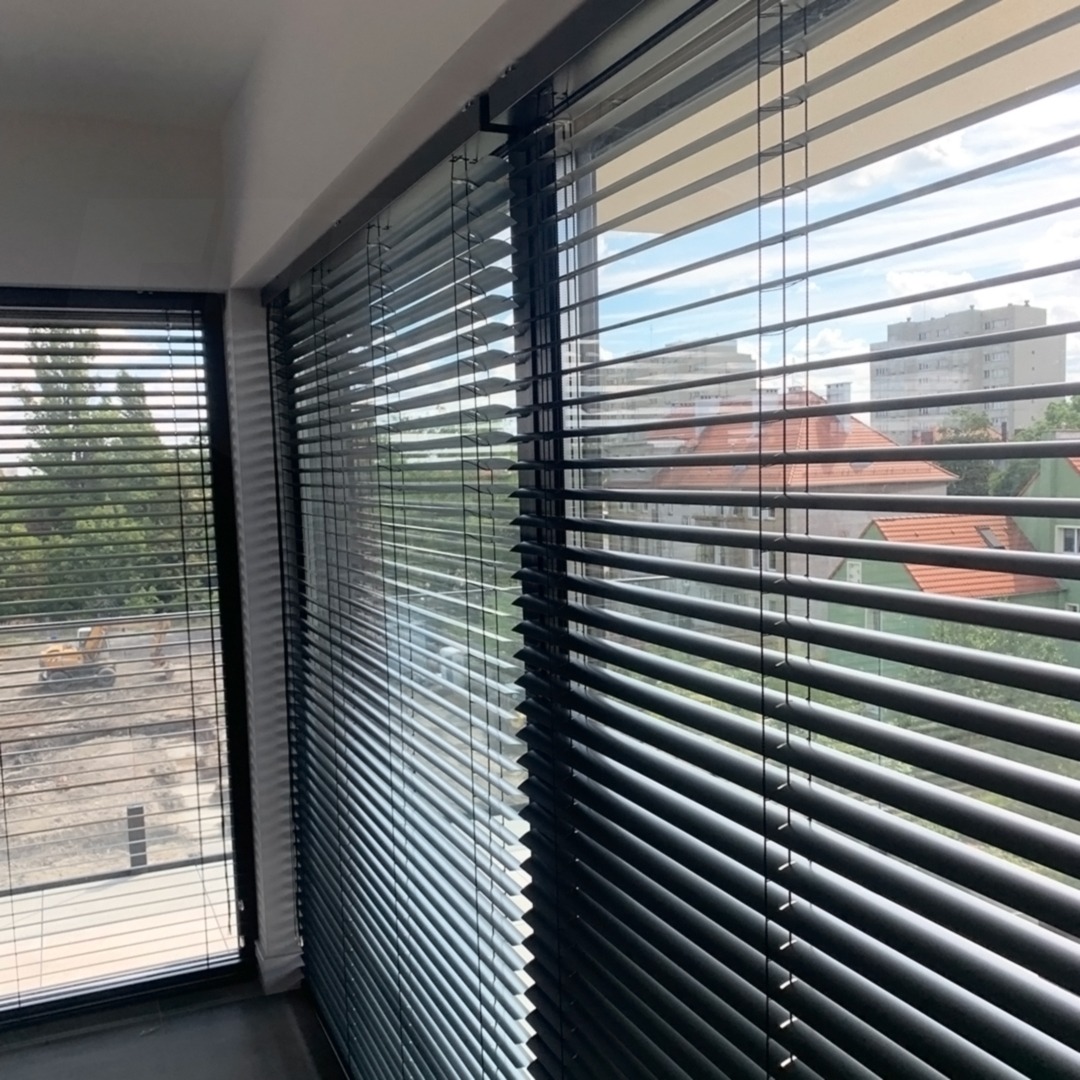 Czarne poziome lamele okienne zamontowane w dwóch oknach, widok na plac budowy i budynki mieszkalne w tle.