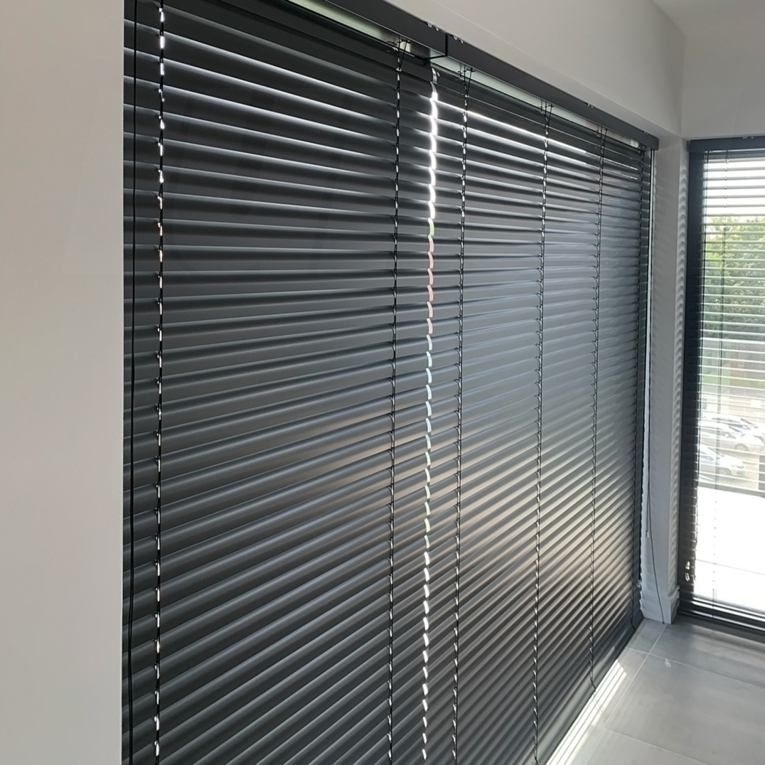 Ciemnoszare poziome osłony okienne z metalowymi listwami, zamontowane w nowoczesnym wnętrzu z jasnymi ścianami i podłogą, wpadające promienie słoneczne.